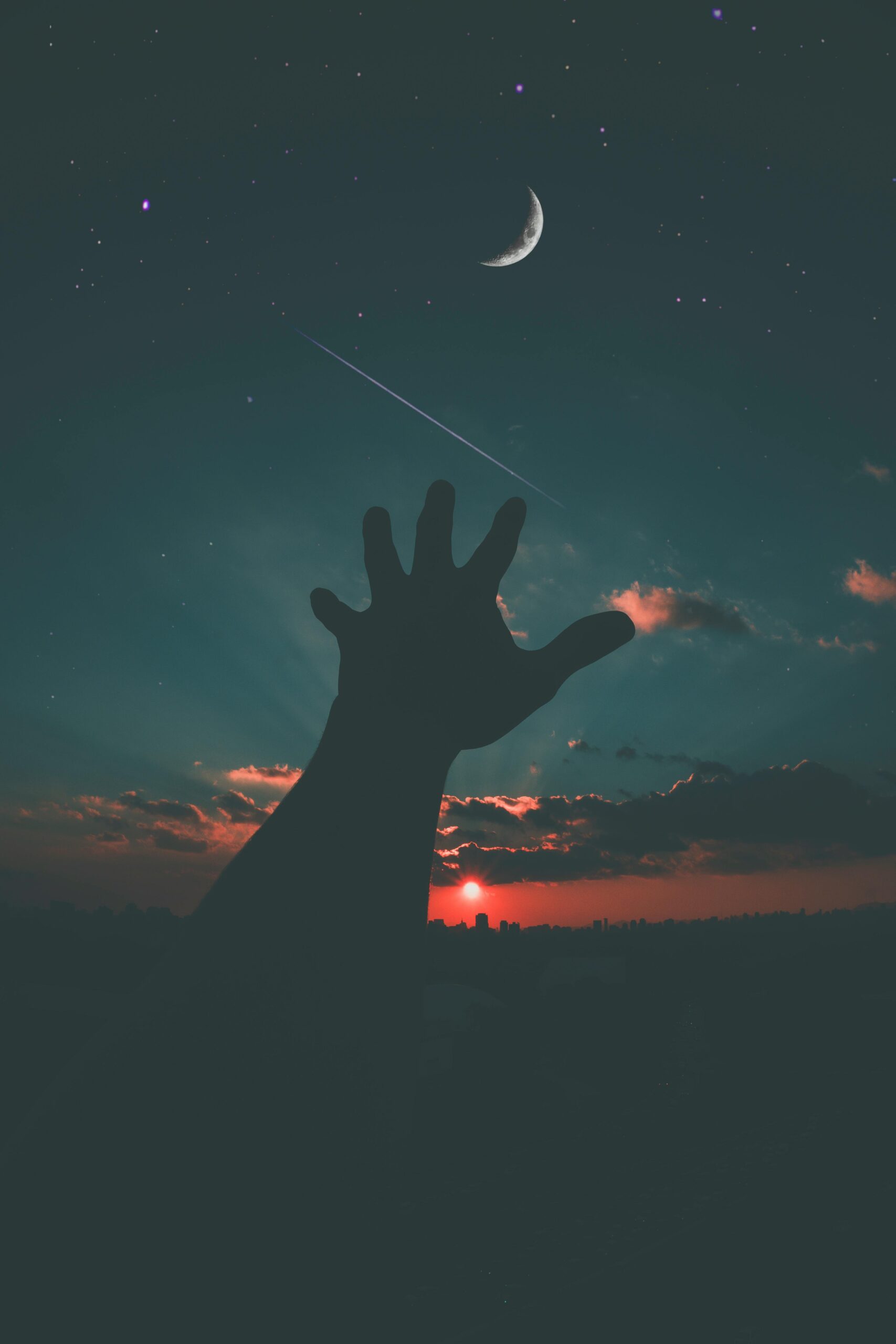 Silhouette of a hand reaching towards the crescent moon and stars in a dramatic dusk sky.