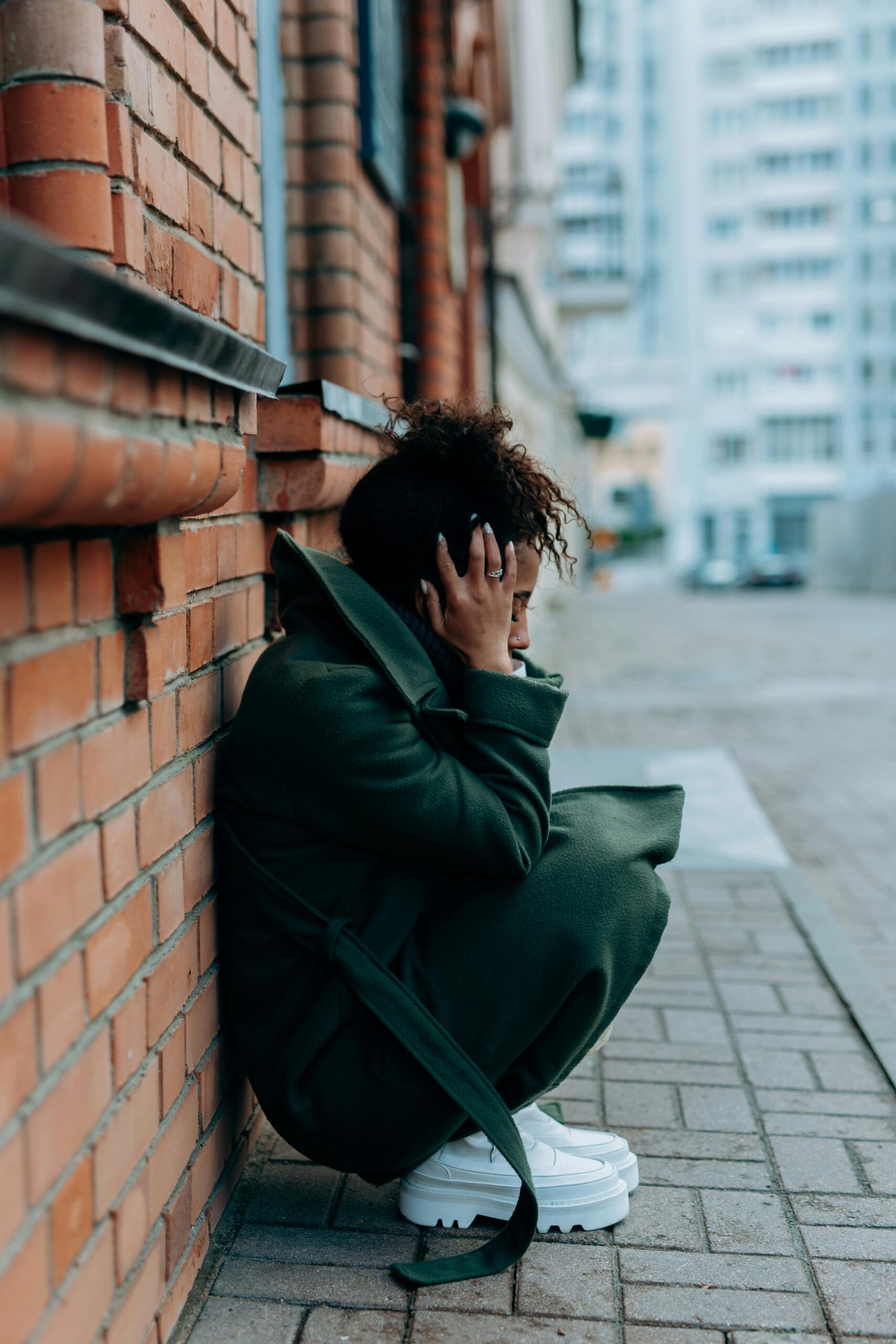 A distressed woman sits alone on a city sidewalk leaning against a brick wall.