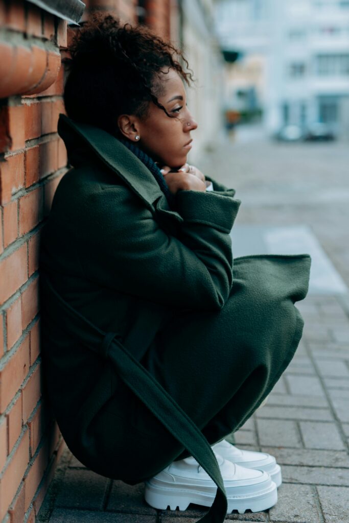 A woman in a trench coat leans against a brick wall, looking contemplative on a city sidewalk.