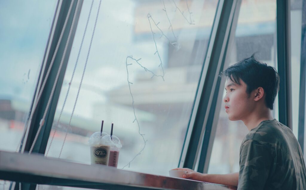 A young man sitting alone, drinking coffee by a large window in a cafe.